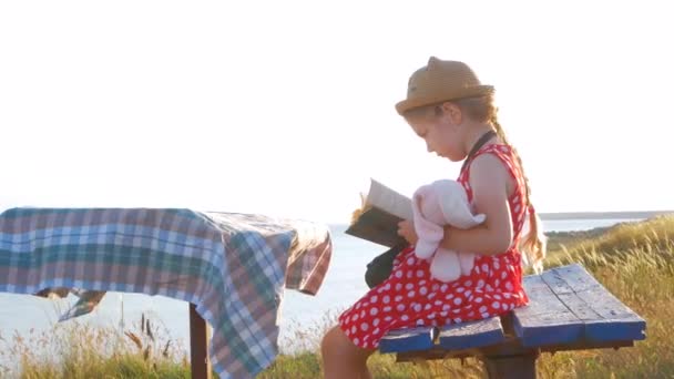 Děvče v slamáku a šaty sedí na lavičce a čte si knihu. Roztomilé dítě s měkkým králík hračka při pohledu na notebook v rukou na mořském přírodním pozadí krajiny. Koncept přátelství. - Záběry, video