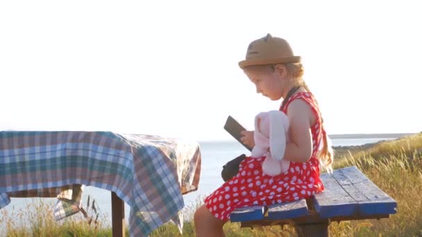 Gyermek lány szalmakalapban és ruhában ül egy régi padon és könyvet olvas. Aranyos gyerek puha nyúl játék nézi notebook kezében tengeri természet lanscape háttér. Barátság fogalma. - Felvétel, videó