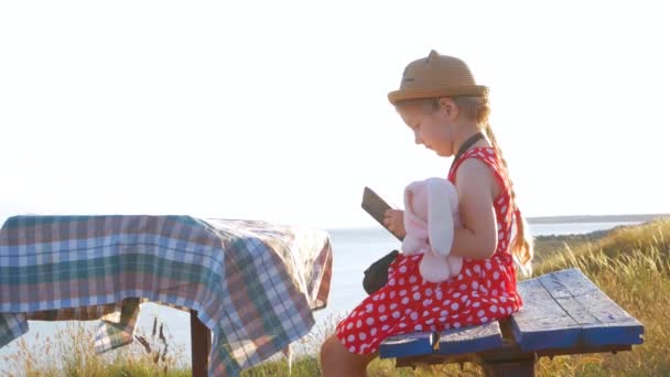 Gyermek lány szalmakalapban és ruhában ül egy régi padon és könyvet olvas. Aranyos gyerek puha nyúl játék nézi notebook kezében tengeri természet lanscape háttér. Barátság fogalma. - Felvétel, videó