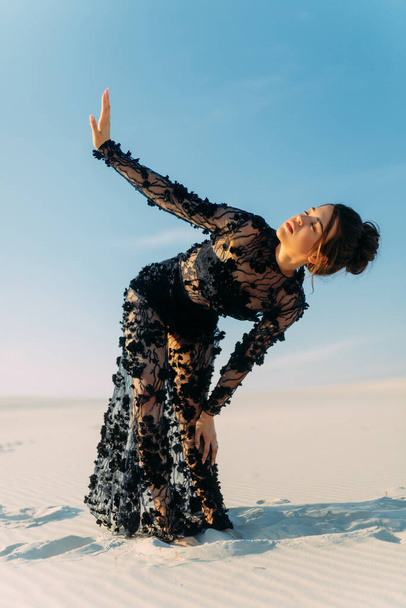 Женщина в кружеве черное платье стоит в пустыне на белом песке и закрывает глаза рукой от солнца на фоне песчаного ландшафта и голубого неба. - Фото, изображение