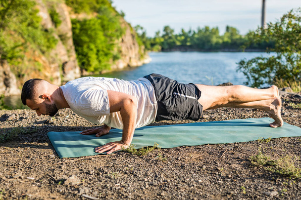 Sabah ve akşam yakışıklı genç bir adam yazın açık havada taş ocağı gölünün kıyısında yoga egzersizi yapıyor. Sağlıklı aktif yaşam tarzı kavramı. - Fotoğraf, Görsel