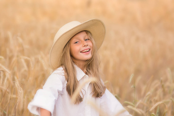 Adorable little girl in a straw hat in wheat field. Child with long blonde wavy hair on countryside landscape with spikelet in hand. Farming agriculture harvesting concept. - 写真・画像