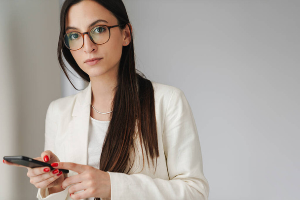 Photo of concentrated businesswoman in eyeglasses using mobile phone while standing indoors - Fotó, kép