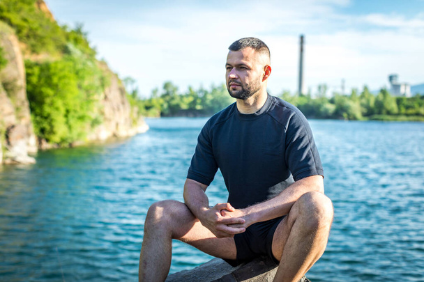 Бородатый молодой человек, сидящий на скалах на фоне карьерного озера. Красивый портрет. - Фото, изображение