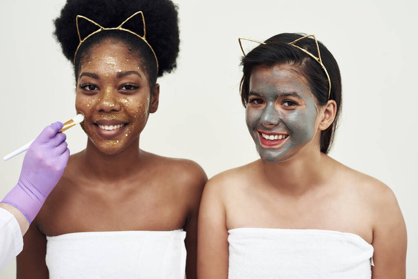 美容師は二人の女性の一人にフェイスマスクを適用します。友情、美しさ、パーソナルケア、化粧品の概念. - 写真・画像