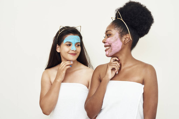 Happy different young women stand in the studio in a towel and laugh, talk among themselves in cosmetic masks. The concept of friendship, beauty, personal care and cosmetics. - Fotoğraf, Görsel