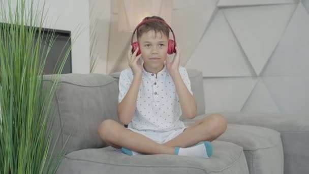 Iloinen afroamerikkalainen poika laittaa kuulokkeet päähän ja siirtyy musiikkiin. Muotokuva iloinen söpö lapsi nauttii harrastus kotona. Onnellinen lapsi istuu sohvalla sisällä ja pitää hauskaa. - Materiaali, video