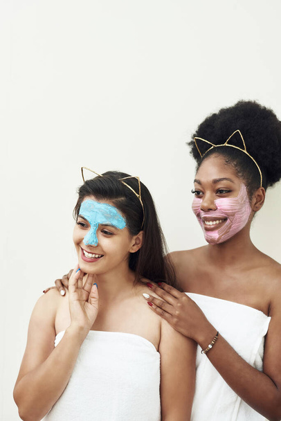 Multimasking. Two different ethnic girls are hugging. The girls' faces are covered with pink and blue face care masks. Beauty face, facial treatment and spa concept. - Photo, Image