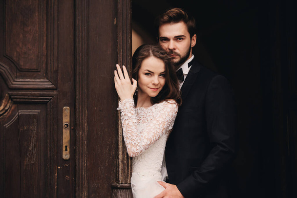 Beautiful newlyweds hugging near the ancient door. Wedding portrait of a stylish groom and a young bride near old house in in a European town. - Photo, Image