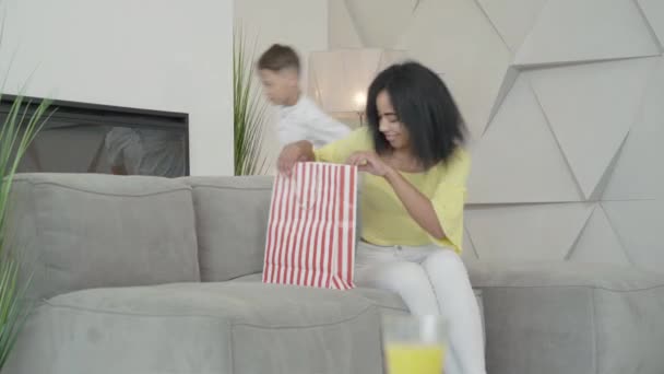 Happy African American woman entering room with shopping bag as son passing by at the background. Portrait of carefree female shopper enjoying purchase at home. Shopaholism concept. - Filmati, video