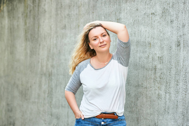 Outdoor portrait of pretty woman posing on grey urban background, wearing white t-shirt with grey sleeves - Photo, Image