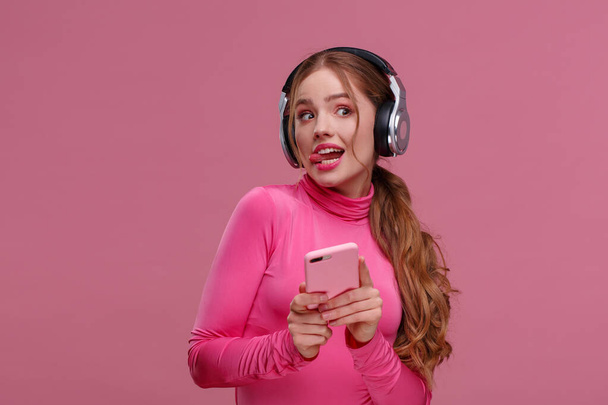 Funny redhead girl holding mobile phone waiting for messages and congratulations from friends. Young smiling woman wearing earphones holding smartphone over pink background. Copy space - Фото, изображение