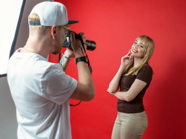Joven modelo hermosa posando a fotógrafo en estudio sobre fondo rojo. Concepto profesional, educativo y tecnológico - Foto, Imagen
