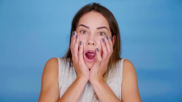 Geschockte Frau öffnet Mund und legt Handflächen auf Wangen vor blauem Hintergrund - Filmmaterial, Video