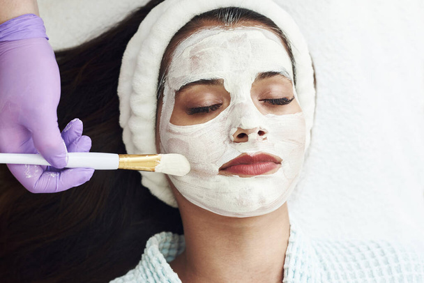 Spa concept. Young woman with nutrient facial mask in beauty salon, close up. The beautician applies a moisturizing mask to the client's face. - Photo, Image