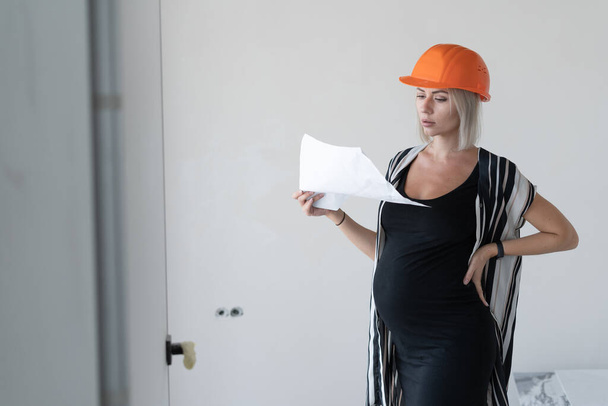 giovane donna incinta che indossa un casco protettivo da costruzione sulla testa. Guarda il foglio di lavoro. Sta contro un muro bianco. - Foto, immagini