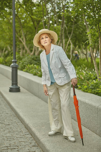 Aged caucasian active and healthy woman about 60 years old is travelling. Elderly tourist in casual cloth and with orange umbrella outdoor. - 写真・画像
