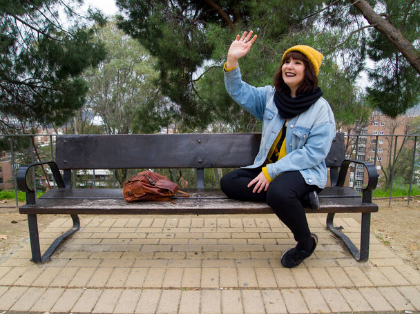 Young woman with yellow woolen cap sitting on a bench waving at someone. Autumn urban concept. - Foto, Imagem