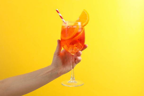 Vrouwelijke hand met glas aperol spritz cocktail op gele achtergrond - Foto, afbeelding