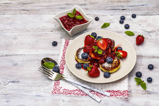Cottage cheese pancakes with fresh berries and strawberry sauce. Healthy breakfast concept. Ripe blueberry, fragrant mint leaves, cutlery. Wooden boards background, copy space - Zdjęcie, obraz