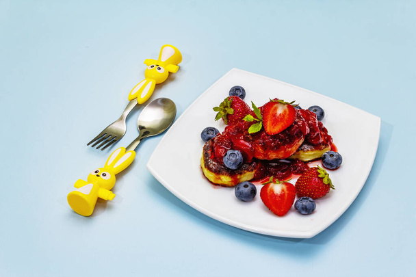 Quark-Pfannkuchen mit frischen Beeren und Erdbeersoße. Gesundes und lustiges Frühstückskonzept für Kinder. Reife Blaubeeren, duftende Minzblätter, Besteck. Sanfter blauer Hintergrund, Kopierraum - Foto, Bild
