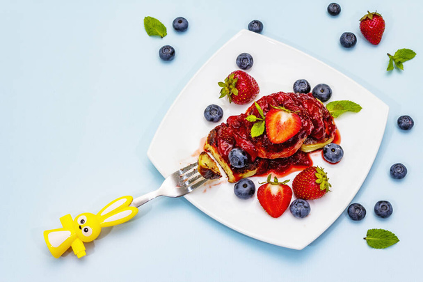 Cottage cheese pancakes with fresh berries and strawberry sauce. Healthy and funny breakfast concept for kids. Ripe blueberry, fragrant mint leaves, cutlery. Gentle blue background, top view - Photo, Image