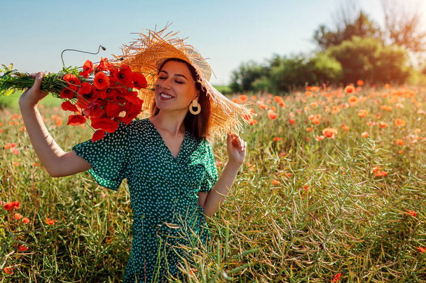 Jeune femme heureuse sentant bouquet de fleurs de coquelicots marchant dans le champ d'été. Fille élégante portant chapeau de soleil de paille. Accessoires pour femmes - Photo, image