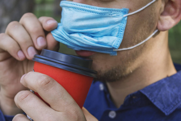 Guy in a medical mask drinks coffee from an orange takeaway cup - Photo, Image