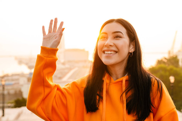 Photo of joyful beautiful asian woman smiling and waving hand while walking outdoors - Foto, imagen