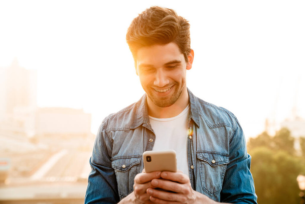 Photo of joyful handsome man smiling and using mobile phone while walking outdoors - Zdjęcie, obraz