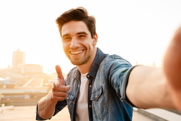 Photo of joyful unshaven young man pointing finger at camera while taking selfie photo outdoors - Photo, Image
