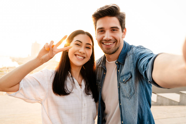 Image of smiling multicultural couple gesturing peace sign and taking selfie photo while walking outdoors - Foto, Bild