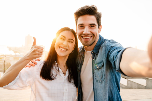 Image of smiling multicultural couple showing thumb up and taking selfie photo while walking outdoors - Foto, Imagem