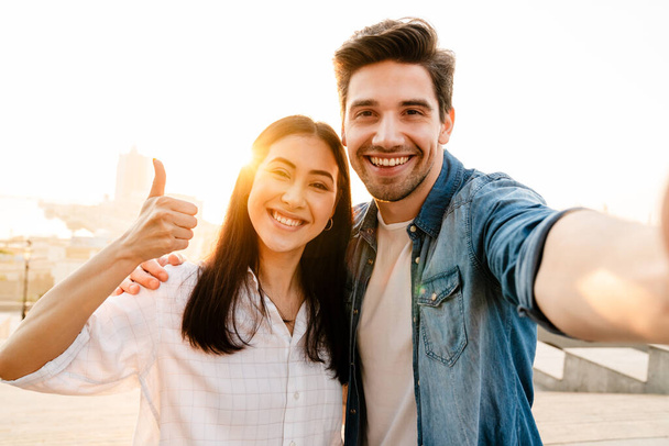 Image of smiling multicultural couple showing thumb up and taking selfie photo while walking outdoors - Foto, immagini