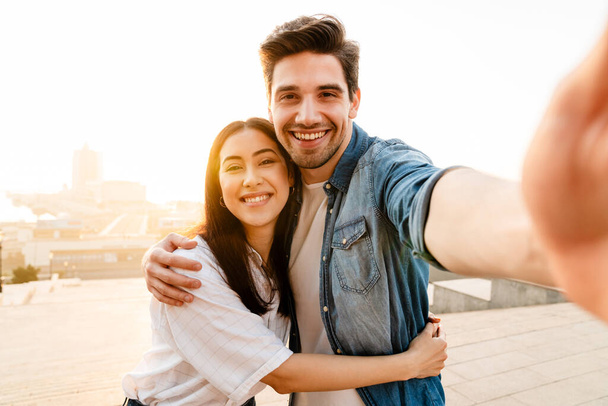Image of smiling multicultural couple hugging and taking selfie photo while walking outdoors - Photo, image