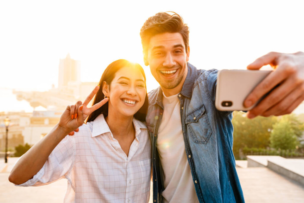 Image of happy multicultural couple gesturing peace sign and taking selfie photo on cellphone outdoors - Foto, Bild