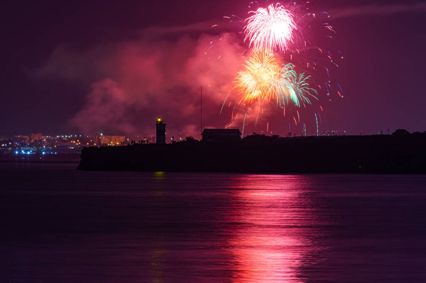Εορταστικός χαιρετισμός πάνω από την πόλη της Σεβαστούπολης την ημέρα του Ναυτικού. Φωτεινές πολύχρωμες λάμψεις πυροτεχνημάτων. Η έννοια της γιορτής. Νυχτερινά γυρίσματα της πόλης. Σιλουέτα του φάρου. - Φωτογραφία, εικόνα