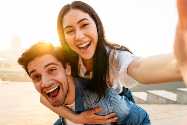 Image of cheerful multicultural couple piggybacking ride and taking selfie photo while walking outdoors - Foto, Imagem