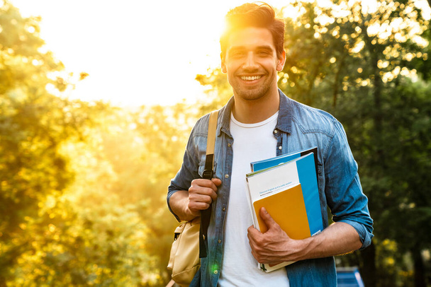 Фото щасливого незахищеного студента з книгами вправ, посміхаючись на камеру під час ходьби в зеленому парку
 - Фото, зображення