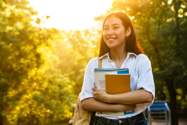 Foto de mujer estudiante asiática feliz con libros de ejercicios sonriendo mientras camina en el parque verde - Foto, Imagen