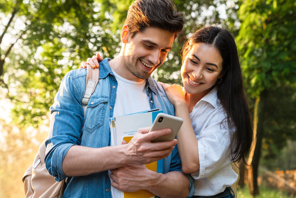 Image of cheerful multicultural student couple using mobile phone and smiling while walking in green park - Foto, afbeelding