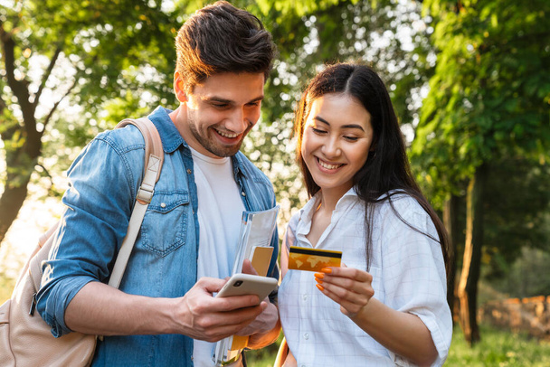 Image of cheerful multicultural student couple using mobile phone and holding credit card while walking in green park - Фото, зображення