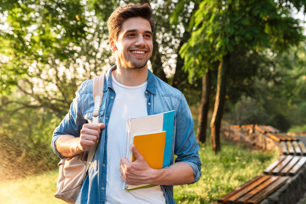 Photo of happy unshaven student man with exercise books smiling while walking in green park - Φωτογραφία, εικόνα