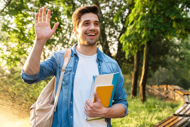 Zdjęcie szczęśliwego studenta z książkami z ćwiczeniami uśmiechniętymi i machającymi ręką podczas spaceru po zielonym parku - Zdjęcie, obraz