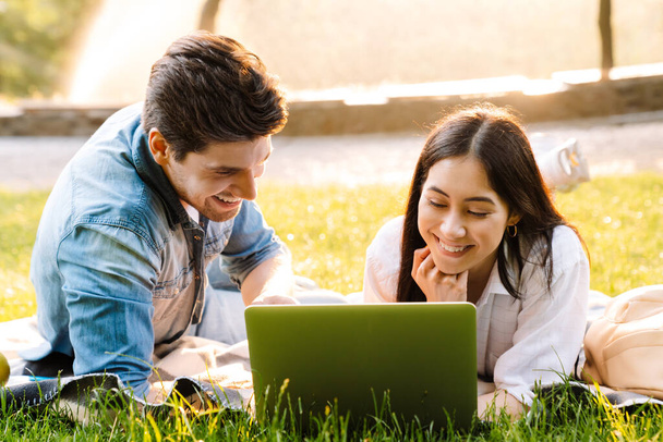 Image of joyful multicultural couple using laptop and smiling while lying on grass in park - Fotoğraf, Görsel