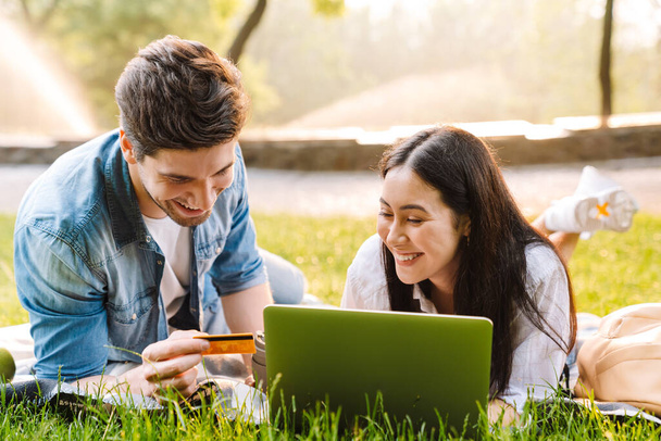 Image of joyful multicultural couple using laptop and holding credit card while lying on grass in park - Φωτογραφία, εικόνα