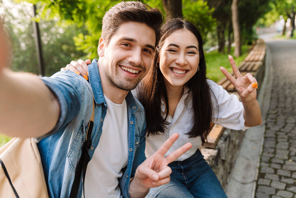 Image of joyful multicultural couple taking selfie photo and gesturing peace sign while sitting on bench in park - Photo, image