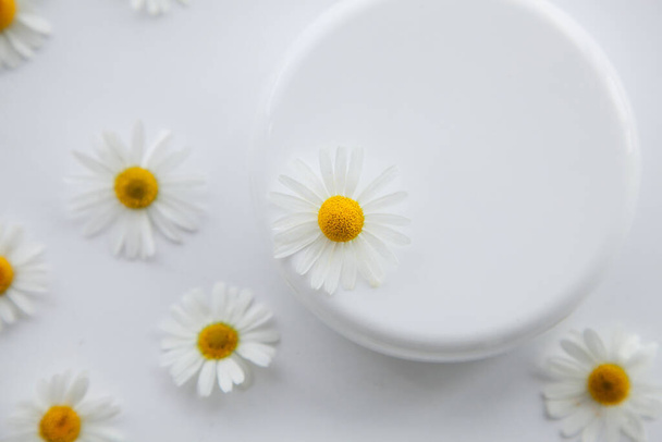 Tarro blanco de crema hidratante y flores de manzanilla sobre un fondo blanco. - Foto, imagen