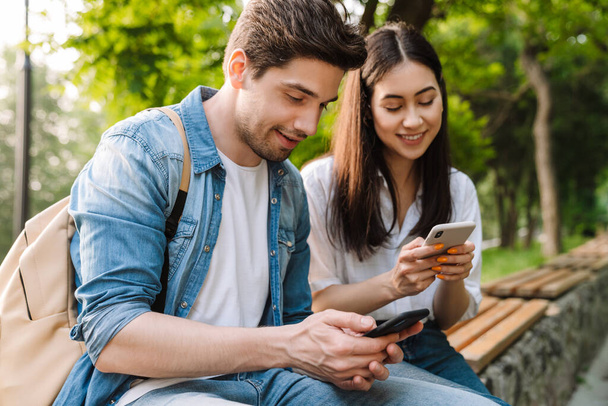 Image of pleased multicultural couple using mobile phones and smiling while sitting on bench in park - Фото, изображение
