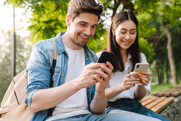 Image of pleased multicultural couple using mobile phones and smiling while sitting on bench in park - Photo, Image